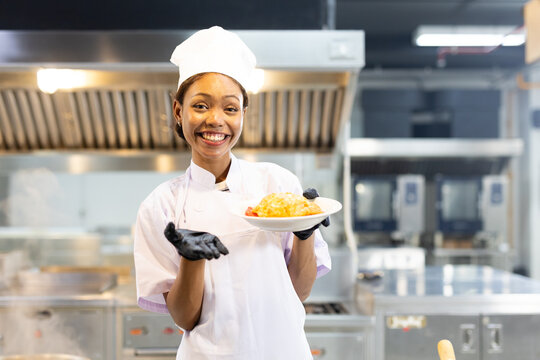Enthusiastic women chef poses with plate dish of food in kitchen. Female wears chef hat and gloves, smiling and giving spread your hands, showing satisfaction in meal has prepared.