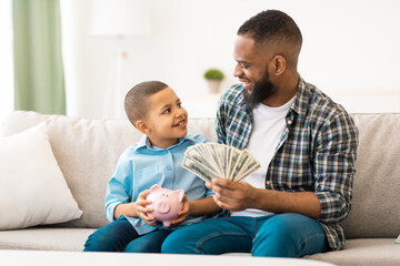 African Father Teaching His Son Budget Planning And Financial Literacy Putting Money Savings In Piggybank Sitting On Couch Indoors. Finances Planning, Banking And Investment Concept. Selective Focus