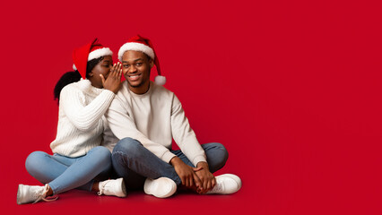 Romantic Black Woman Whispering To Boyfriend's Ear, Sharing Christmas Secret With Her Lover, Smiling African Couple Wearing Santa Hats, Having Fun Together While Sitting On Orange Background