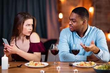 Black Man Angry To His Girlfriend Texting On Smartphone During Date In Restaurant, Young Guy Blaming His Lover For Cheating And Infidelity. Multiracial Couple Having Problems In Relationship