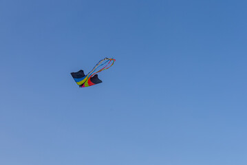 A bright, multi-colored kite with long, flowing tails is captured mid-flight against a clear, deep blue sky. 