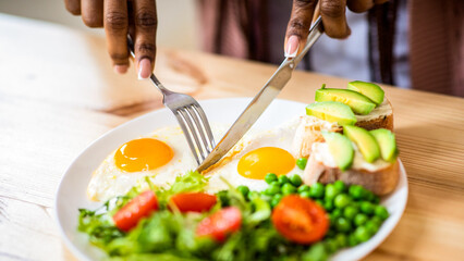 Unrecognizable black female using fork and knife while eating tasty breakfast in kitchen, african american woman enjoying morning meal, having plate with fresh salad, eggs and sandwiches, closeup