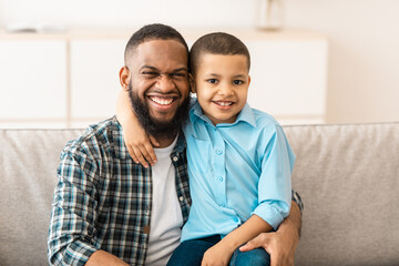 Father's Day. Happy African American Daddy And His Little Son Hugging Posing Together Embracing And Smiling To Camera Sitting On Couch At Home. Portrait Of Happy Dad And Kid Boy. Fatherhood Concept