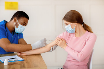 Fototapeta premium Millennial woman in medical mask scared of coronavirus vaccine injection, looking at syringe with fear at hospital. Doctor making covid-19 vaccination to young lady. Immunization against global virus