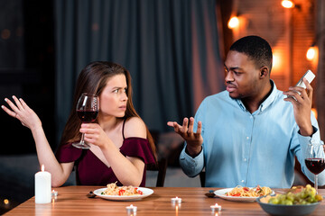 Young Woman Complaining About Her Black Boyfriend Talking On Cellphone During Romantic Date In Restaurant, Feeling Annoyed And Bored. Interracial Couple Having Problems In Relationship, Free Space