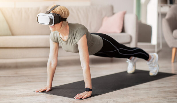 Exercising with virtual reality concept. Mature woman in VR headset standing in plank pose at home. Strong senior lady using modern technologies for domestic yoga workout