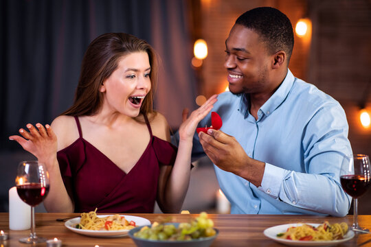 Proposal Surprise. Romantic young black man proposing marriage to his beautiful girlfriend at date in restaurant, holding box with diamond ring in hand, joyful woman raising hands in happy excitiment