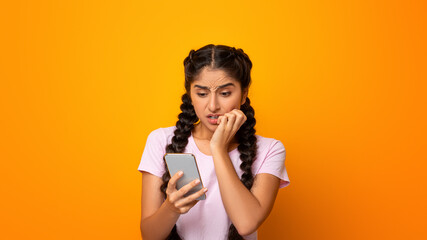 Portrait of anxious scared indian young woman looking at mobile phone seeing bad news, photos or message. Worried lady biting nails isolated on yellow studio background. Human reaction, expression