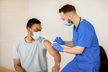 Fototapeta premium Doctor in face mask giving covid-19 vaccine injection to black male patient during vaccination campaign at clinic. Young African-American man getting immunized against infectious disease at hospital