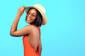 Fototapeta premium Summer fun. Portrait of cool black lady in stylish sunglasses, straw hat and bikini posing over blue background. Fashionable African American woman in trendy swimwear smiling at camera