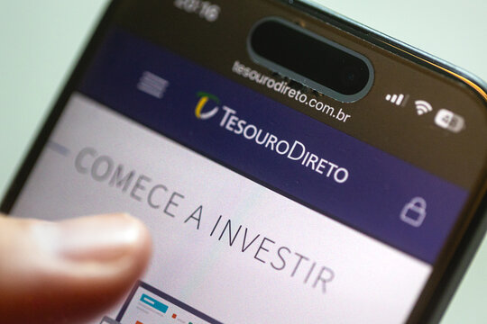 Um homem segurando um smartphone e mostrando o site do Tesouro Direito. Investimento, Renda fixa, Governo Brasileiro, compra e venda de t&iacute;tulos p&uacute;blicos.