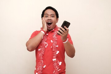 Happy Indonesian man celebrating Independence Day with red and white accessories, holding national flag and smartphone in excitement.