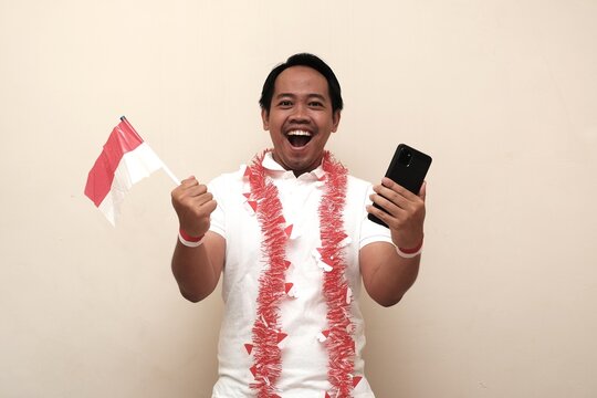 Happy Indonesian man celebrating Independence Day with red and white accessories, holding national flag and smartphone in excitement. - Powered by Adobe