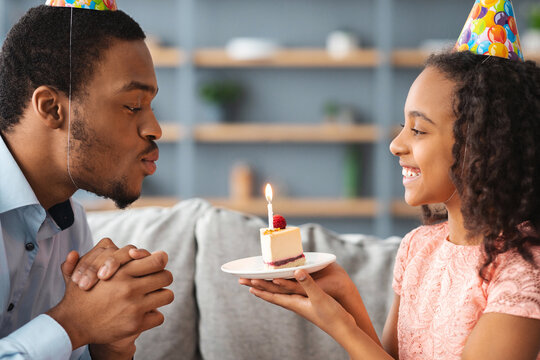 Young black man blowing candle on birthday cake in his smiling cute little daughter hands, closeup portrait. Loving african american family having birthday celebration at home, wearing party hats - Powered by Adobe