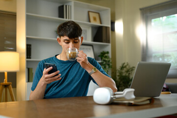 Young man drinking coffee while checking smartphone at home office desk with laptop