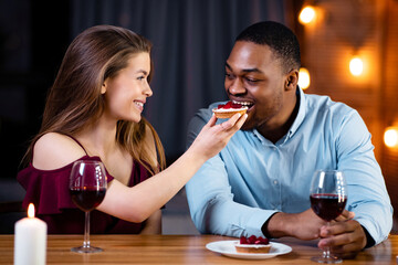 Romantic interracial couple having dinner date in restaurant, feeding each other with desserts and drinking wine, black man and while woman having fun while celebrating valentine's day together