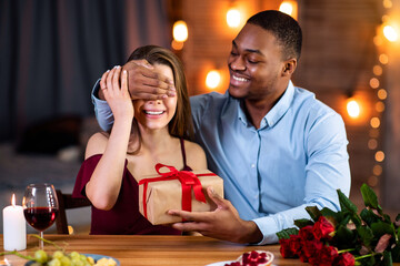 Loving African American Man Covering White Girlfriend's Eyes And Presenting Wrapped Gift Box At Romantic Date In Restaurant. Happy Young Interracial Couple Celebrating Valentine's Day Together
