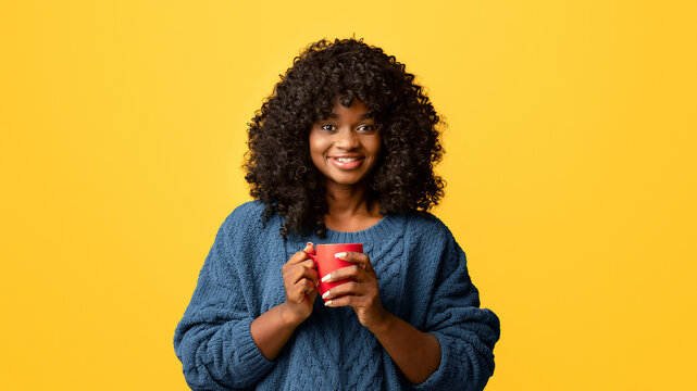 Smiling young black woman in casual knitted sweater holding red mug with hot drink, drinking tea or coffee, copy space, yellow studio background, panorama. Millennials lifestyle, candid photos - Powered by Adobe