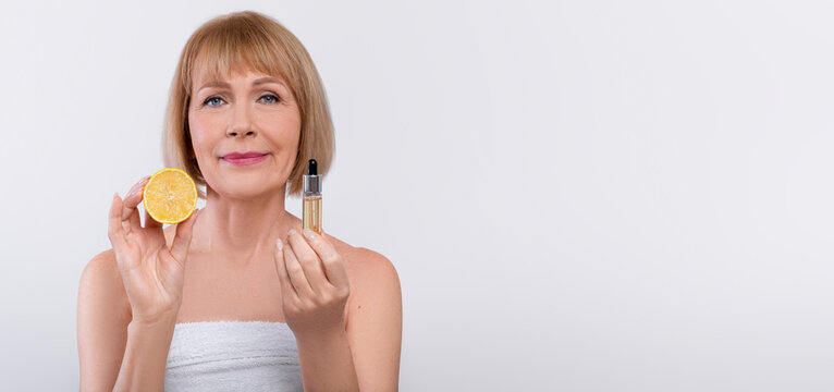 Beautiful mature woman showing bottle with vitamin C serum on light studio background, copy space. Sensual senior lady holding anti-aging cosmetic product, closeup