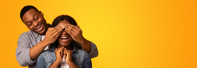Fototapeta premium Cheerful black guy surprising his excited girlfriend, covering her eyes from the back and telling guess who, joyful african american lovers standing isolated over yellow background, free space
