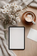 Flat lay of a blank white iPad Pro mockup on a light wooden table next to a cappuccino, dried flowers and a greeting card