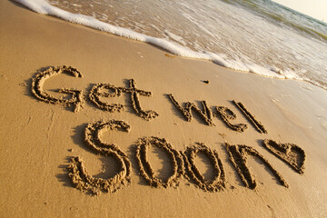 Fototapeta premium Get Well Soon Handwritten in Beach Sand with Minimalist Heart and Cross Illustration and Ocean Foam in Caring Vertical Coastal Scene