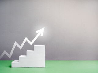 Glowing white upward zigzag arrow rise on stair blocks as business graph steps on green table, grey background with copy space. Business growth process, growing economy, economic improvement concept.
