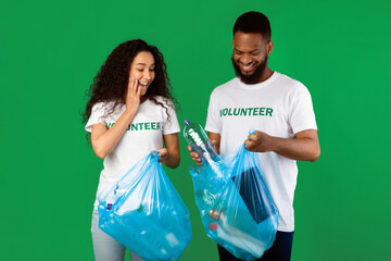 Sort Your Waste. Two Multiracial Volunteers Couple Putting Used Plastic Bottle To Garbage Bag Posing Standing In Studio Over Green Background. Keep Environment Clean, Ecology Concept