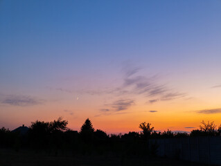 A sunset with the moon in the sky and trees in the foreground