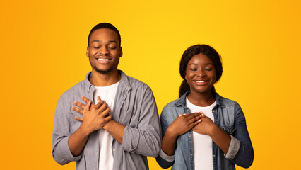 With Faith In Heart. Black couple praying with hands on chest and closed eyes, blessed african american man and woman standing isolated over yellow background, cherishing hope for best