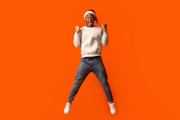 Overjoyed Black Guy In Santa Hat Celebrating Success, Jumping Over Orange Background With Raised Fists, Euphoris African American Man Enjoying Christmas Offers And Seasonal Sales, Full Length Shot