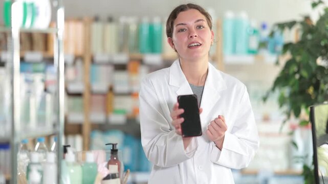 Friendly skilled young girl pharmacist standing behind counter, demonstrating and offering mobile application for smartphone in modern pharmacy