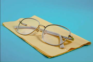 A pair of stylish gold-framed eyeglasses with clear lenses resting on a yellow cleaning cloth against a blue background. Sleek and practical for vision and fashion themes.