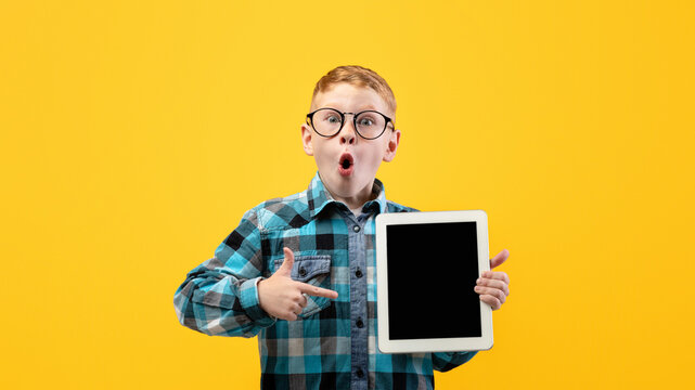 Amazed schoolboy in glasses pointing at empty digital tablet screen, recommending cool educational or entertaining website or application, mockup, yellow studio background, copy space