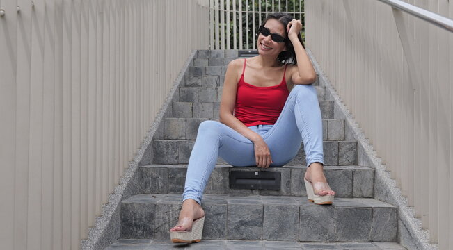 Confident Asian woman in a red tank top and jeans sits casually on stone steps, smiling with a relaxed urban attitude - Powered by Adobe