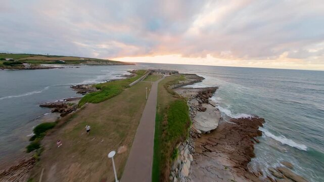 Smooth flight following a path along a small peninsula with a building at the end, facing the sea where the sun is setting and waves gently roll in. Natural, warm light of sunset.
