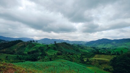 Obraz premium Green mountain and valley landscape in rural Laos with cloudy sky. The dramatic sky above is filled with beautiful grey and white clouds, creating a serene, peaceful, and majestic atmosphere.
