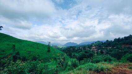 Naklejka premium Green mountain and valley landscape in rural Laos with cloudy sky. The dramatic sky above is filled with beautiful grey and white clouds, creating a serene, peaceful, and majestic atmosphere.