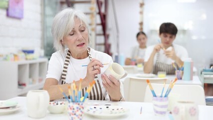Interested elderly woman attending pottery class, enthusiastically painting on ceramic cup, creating unique custom design. Creative hobby concept