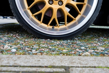 Close-up view of a gold alloy car wheel with a low-profile tire, showing the rim details and valve stem, parked on a stone-paved surface with grass patches.