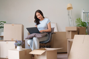Fototapeta premium Asian adult woman actively manages new home relocation using laptop computer for planning and organizing amidst moving boxes in a modern apartment