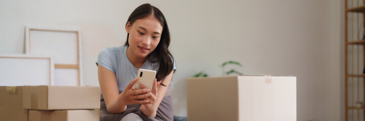 Asian adult woman uses smartphone managing home relocation organizing new apartment surrounded boxes © snowing12