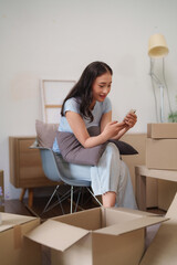 Asian adult woman taking break new apartment looking phone surrounded moving boxes planning relocation future home life