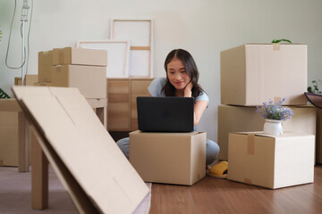 Young Asian woman uses laptop managing moving new apartment surrounded cardboard boxes planning home setup remote work