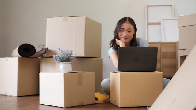Asian young adult woman uses laptop computer among moving boxes planning relocation settling into new apartment new house journey home setup