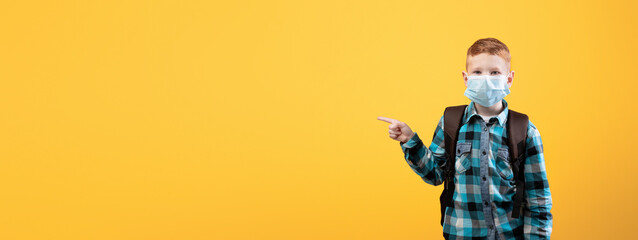 Schoolboy in protective face mask pointing at copy space over yellow studio background. Redhead schoolboy with backpack wearing face mask, using protection against coronavirus while going to school