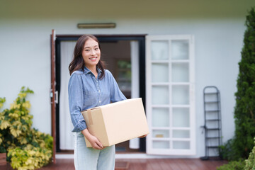 Smiling young adult Asian woman carefully carries cardboard box new home doorstep happy...