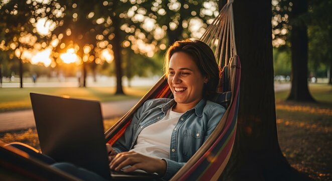 Joyful woman relaxing in a hammock using laptop computer under the sunlight with green park background.