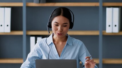 A young Asian woman in a business setting smiles while using her laptop and headset to participate in an online meeting. The modern office features wooden shelves and organization. - Powered by Adobe