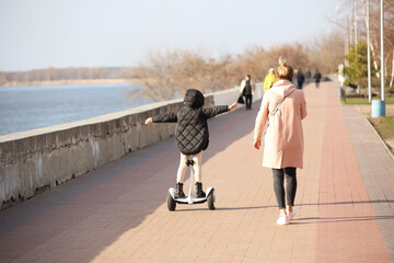 A young mother and daughter walk along the promenade. A girl riding a park on a gyro scooter. Modern electric ecological mode of transport. Ecology in the city. Activity of children. Sports for health